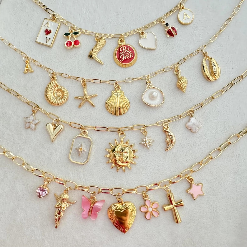 May include: Gold-toned charm necklaces with various pendants, including a playing card, cherries, a banana, a heart, a ladybug, and a seashell. Other charms include a starfish, a sun, a moon, and a cross. The necklaces are displayed on a white surface.
