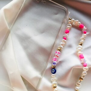 May include: A clear phone case and a beaded phone charm. The charm features white pearls, gold beads, pink star-shaped beads, and letter beads spelling "SUNNY". An evil eye bead is also included.