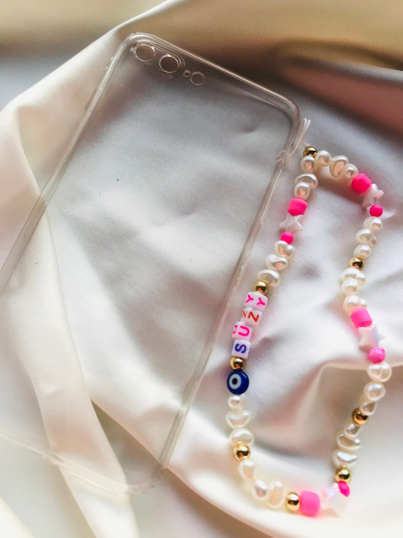 May include: A clear phone case and a beaded phone charm. The charm features white pearls, gold beads, pink star-shaped beads, and letter beads spelling "SUNNY". An evil eye bead is also included.