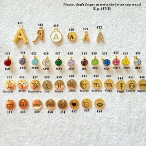 May include: A collection of gold-toned metal charms arranged in rows, featuring letters, birthstones, zodiac symbols, and icons. Each charm is labeled with a unique identification number from 417 to 454, with text indicating months and signs.