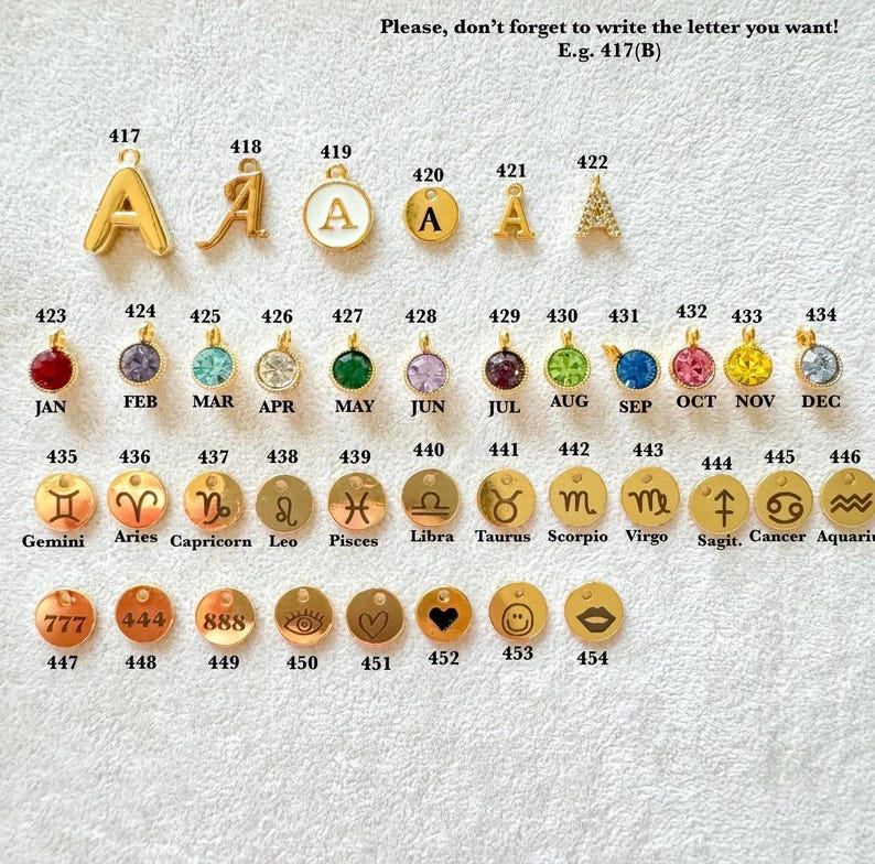 May include: A collection of gold-toned metal charms arranged in rows, featuring letters, birthstones, zodiac symbols, and icons. Each charm is labeled with a unique identification number from 417 to 454, with text indicating months and signs.