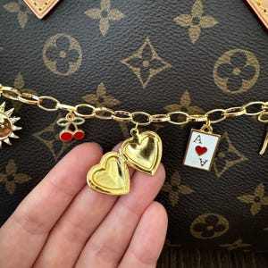 May include: A gold-toned chain bracelet featuring multiple charms including a sun, a pair of red cherries, an open heart-shaped locket, and an ace of hearts playing card charm, set against a brown background with a repeating floral pattern.