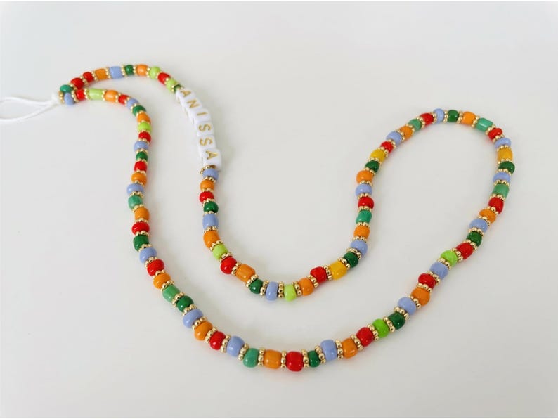 May include: A colorful beaded lanyard with the name "ANISSA" spelled out in white letter beads. The lanyard features a variety of beads in red, orange, yellow, green, and blue, with gold-colored spacers. A white string is attached.