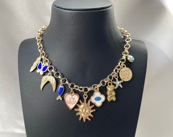 May include: Gold-toned charm necklace with various pendants, including wings, a fish, a heart with "I'M GOOD" text, a sun, a four-leaf clover, a teddy bear, and a starfish. The charms are in various colors, including blue, white, and gold.