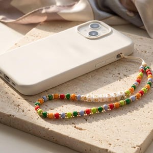 May include: A white phone case with a colorful beaded phone strap. The strap features the name "ANISSA" spelled out in white letter beads, interspersed with red, orange, green, and blue beads. The phone rests on a textured surface.