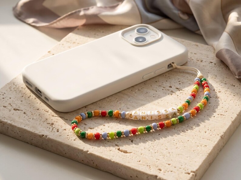 May include: A white phone case with a colorful beaded phone strap. The strap features the name "ANISSA" spelled out in white letter beads, interspersed with red, orange, green, and blue beads. The phone rests on a textured surface.