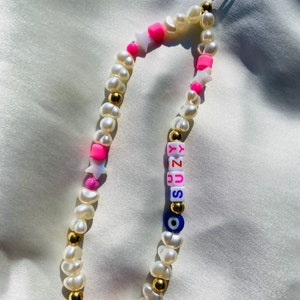 May include: A phone charm made of white pearls, gold beads, and bright pink elements. The charm includes star-shaped beads, the name "SUZY" in colourful letter beads, and an evil eye bead.