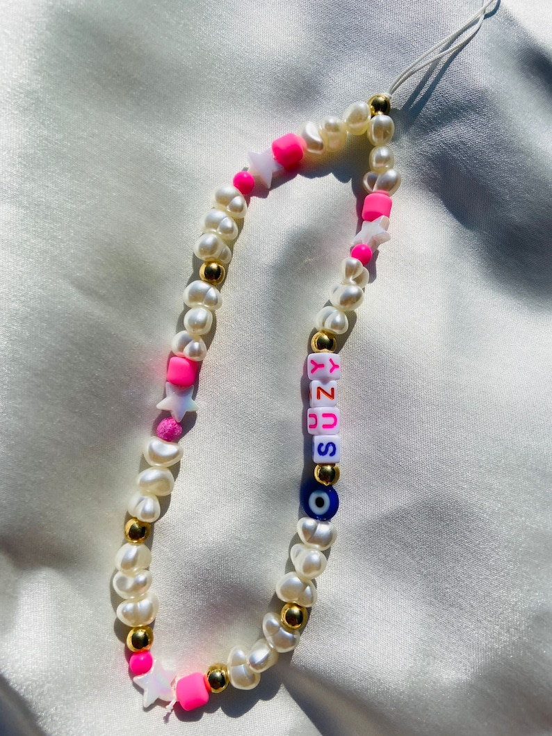 May include: A phone charm made of white pearls, gold beads, and bright pink elements. The charm includes star-shaped beads, the name "SUZY" in colourful letter beads, and an evil eye bead.