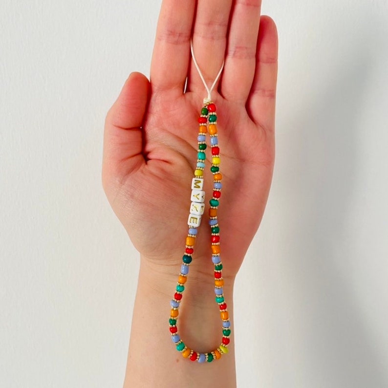 May include: A beaded phone charm with the word "LOVE" spelled out in white letter beads. The charm features a variety of colorful beads, including red, yellow, green, blue, and orange, strung together on a white cord.