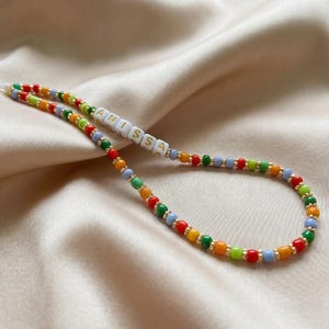 May include: A colorful beaded phone charm with the name "ANISSA" spelled out in white letter beads. The charm features a variety of small beads in red, orange, yellow, green, and blue, strung together with gold-colored accents. The charm is on a white cord.