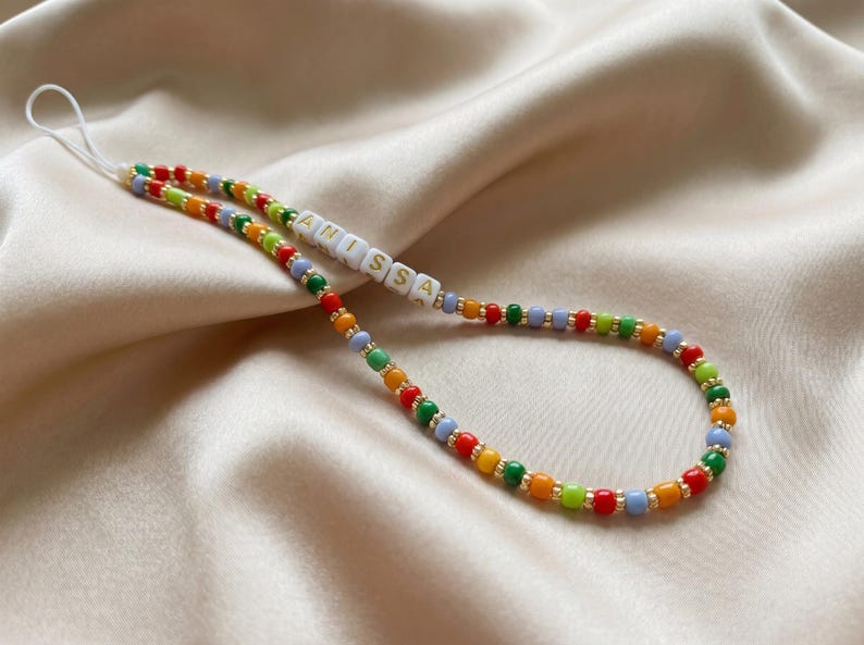 May include: A colorful beaded phone charm with the name "ANISSA" spelled out in white letter beads. The charm features a variety of small beads in red, orange, yellow, green, and blue, strung together with gold-colored accents. The charm is on a white cord.