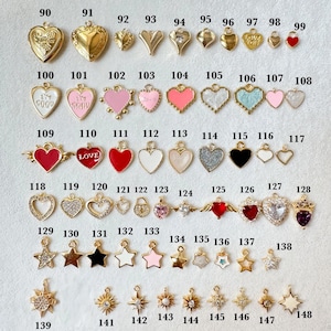 K&ouml;nnte beinhalten: Eine Sammlung goldfarbener und farbiger Charms in verschiedenen Formen, darunter Herzen, Sterne und Medaillons. Einige Charms weisen Glitzer, Emaille und Text auf. Die Charms sind f&uuml;r die Schmuckherstellung und das Basteln konzipiert.