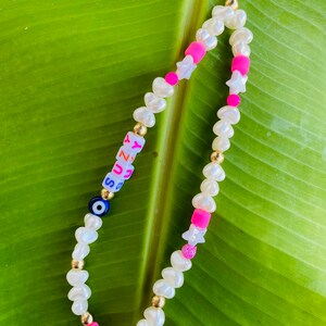 May include: A phone charm with white pearl-shaped beads, gold-coloured beads, and pink and white star-shaped beads. The charm includes a blue evil eye bead and letter beads spelling "SUNNY".