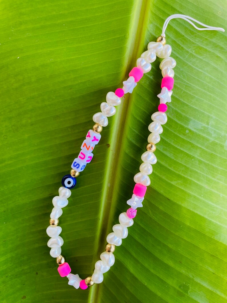 May include: A phone charm with white pearl-shaped beads, gold-coloured beads, and pink and white star-shaped beads. The charm includes a blue evil eye bead and letter beads spelling "SUNNY".
