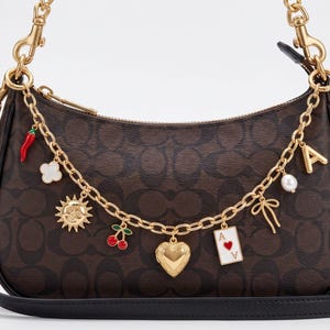 May include: A brown monogram shoulder bag featuring a gold-tone chain strap adorned with various charms, including a red chili pepper, a white flower, a sun, cherries, a heart locket, an ace of hearts playing card, a bow, a pearl, and the letter A.