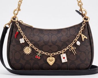 May include: A brown monogram shoulder bag featuring a gold-tone chain strap adorned with various charms, including a red chili pepper, a white flower, a sun, cherries, a heart locket, an ace of hearts playing card, a bow, a pearl, and the letter A.