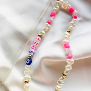 May include: A phone charm made of white pearl beads, gold-coloured accents, and pink and white star-shaped beads. The charm spells out "SUZY" with colourful letter beads and includes a blue evil eye bead.