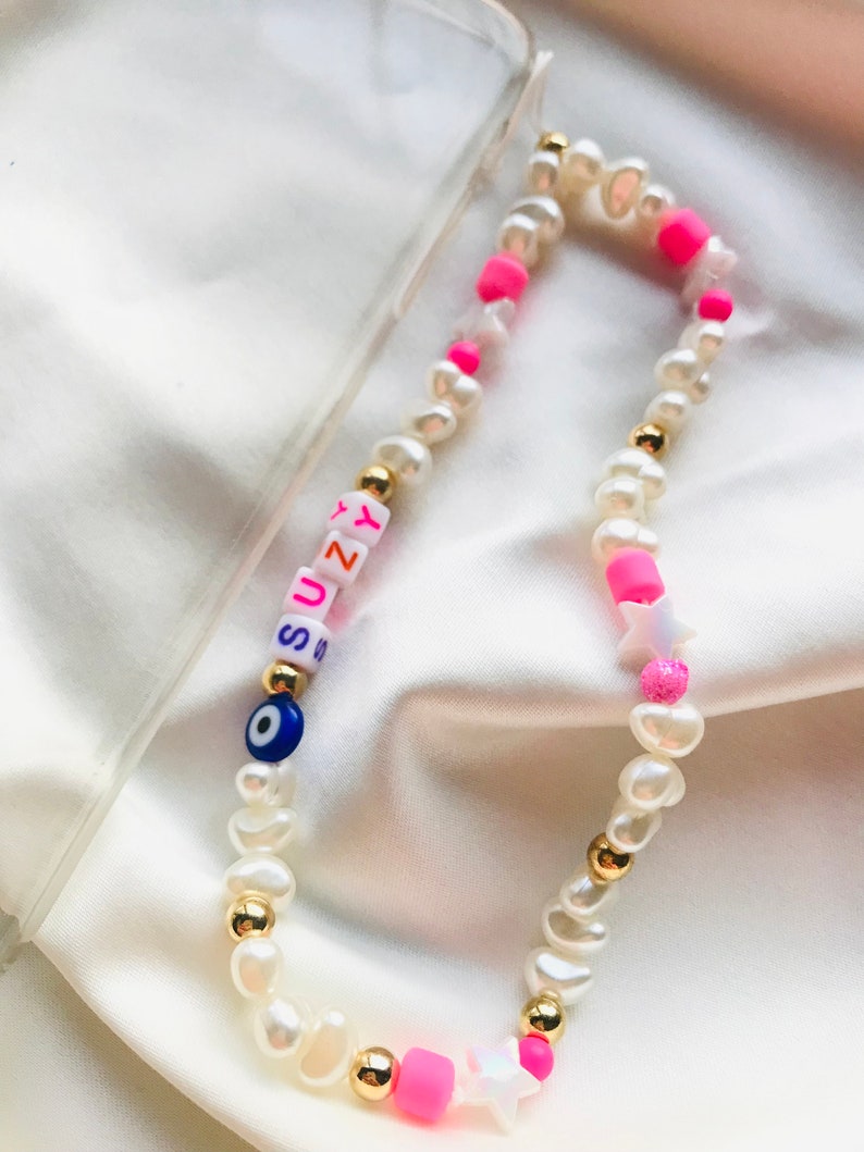 May include: A phone charm made of white pearl beads, gold-coloured accents, and pink and white star-shaped beads. The charm spells out "SUZY" with colourful letter beads and includes a blue evil eye bead.