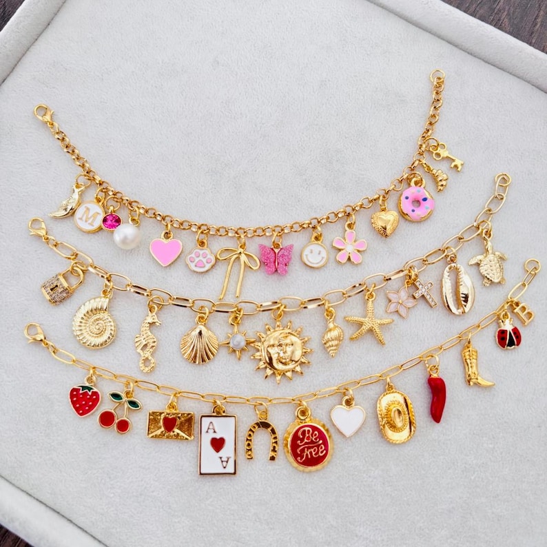 May include: Three gold-colored charm bracelets with various pendants. Charms include a letter "M", a pink heart, a paw print, a butterfly, a smiley face, a flower, a key, a donut, a lock, a seashell, a seahorse, a sun, a starfish, a ladybug, a strawberry, cherries, a playing card, a horseshoe, and a heart.