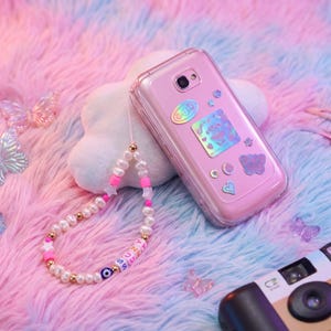 May include: A pink phone case with decorative stickers, a pearl beaded phone charm with the name "SUZY" and a blue evil eye bead. The phone and accessories are on a pink and blue fluffy surface with butterfly decorations.