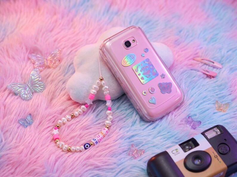 May include: A pink phone case with decorative stickers, a pearl beaded phone charm with the name "SUZY" and a blue evil eye bead. The phone and accessories are on a pink and blue fluffy surface with butterfly decorations.