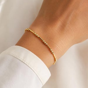May include: A delicate gold bracelet featuring a repeating pattern of small round beads and thin cylindrical bars. The jewelry piece is constructed with a sequence of metallic spheres and straight segments, creating a minimalist and refined appearance.