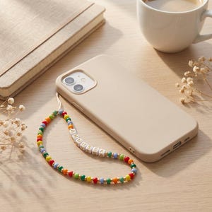 May include: A beige phone case with a colorful beaded phone strap. The strap features a variety of colored beads and white letter beads spelling "ANNUSSA". A notebook and a cup of coffee are in the background.