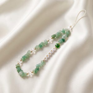 May include: A green gemstone phone charm with the word "KARMA" spelled out in white beads. The charm features a string of green and clear stones, gold beads, and an evil eye bead. The charm is on a white background.