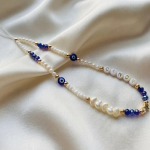 May include: A beaded phone charm with white pearls, blue and gold details, and the name "CINDY" in white letter beads. It features blue evil eye beads and a gold star charm. The charm is designed for a phone.