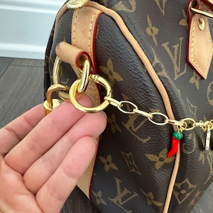 May include: A brown handbag with a repeating monogram pattern, featuring light tan leather trim, gold-tone metal hardware, and a decorative gold chain attachment adorned with a small red and green chili pepper charm.