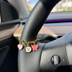 Op de afbeelding: Een goudkleurige ketting met drie bungelende bedels: een lichtroze hartje, een ronde witte bedel met de zwarte letter E en een glinsterende roze vlinder, bevestigd rondom een zwart stuurwiel van een auto.