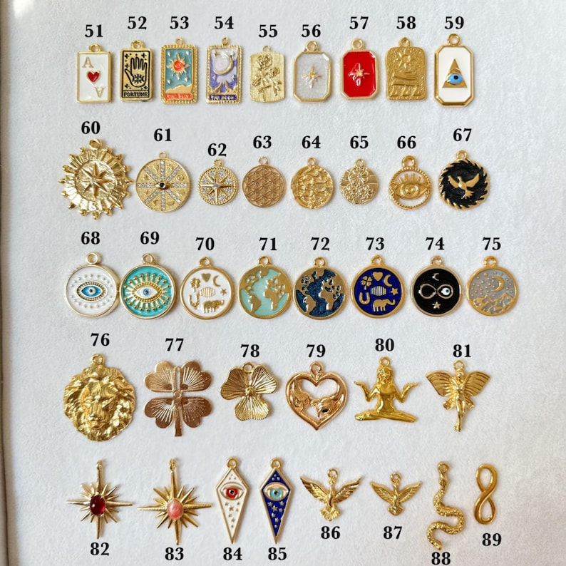 May include: A collection of gold-toned pendants with various designs, including celestial motifs, symbols, and geometric patterns. Some pendants feature enamel detailing in colors like blue, red, and white. The pendants are arranged on a light-colored surface.