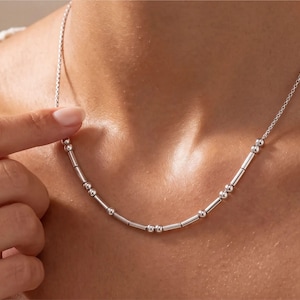 May include: A silver-toned necklace featuring a delicate chain interspersed with a series of small, polished spherical beads and elongated cylindrical metal bars arranged in a pattern that resembles a sequence of Morse code symbols.