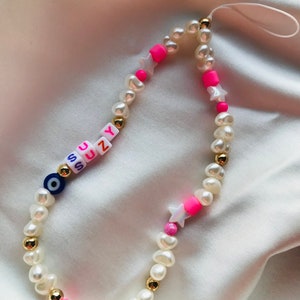 May include: A phone charm made of white pearl beads, gold beads, and pink accents. The charm spells out "SUZY" with letter beads and includes star-shaped beads and a blue evil eye bead.