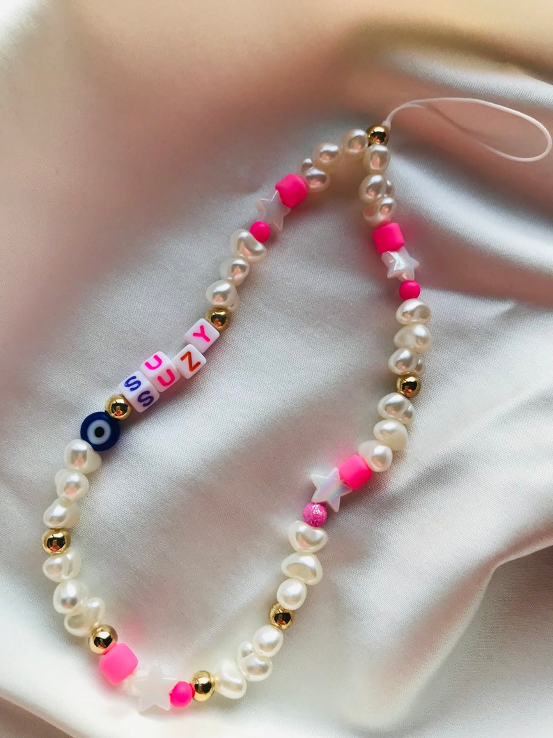May include: A phone charm made of white pearl beads, gold beads, and pink accents. The charm spells out "SUZY" with letter beads and includes star-shaped beads and a blue evil eye bead.
