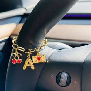 May include: Gold charm bracelet with a cherry, the letter "A", and an envelope with a heart. The charms hang from a chunky chain. The bracelet is shown on a black steering wheel.