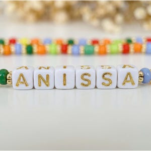 May include: A colorful beaded bracelet spelling "ANISSA" with gold lettering on white cube beads. The bracelet features a variety of beads in red, yellow, green, orange, and blue, with gold accents. The background is blurred, highlighting the bracelet.