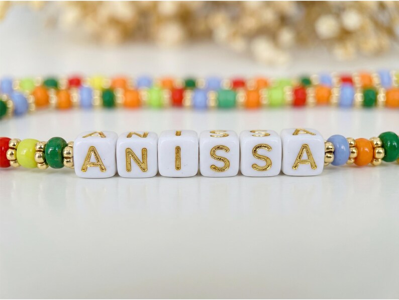 May include: A colorful beaded bracelet spelling "ANISSA" with gold lettering on white cube beads. The bracelet features a variety of beads in red, yellow, green, orange, and blue, with gold accents. The background is blurred, highlighting the bracelet.