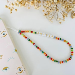 May include: A beaded phone strap with the name "ANISSA" spelled out in white letter beads. The strap features a colorful pattern of red, orange, green, and blue beads. A pink phone case with eye designs is visible in the background.