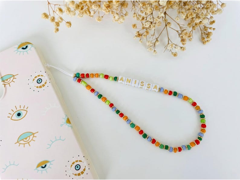 May include: A beaded phone strap with the name "ANISSA" spelled out in white letter beads. The strap features a colorful pattern of red, orange, green, and blue beads. A pink phone case with eye designs is visible in the background.