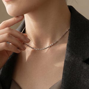 May include: A silver-toned necklace featuring a delicate chain adorned with a repeating pattern of small metallic spheres and short cylindrical bars. The necklace rests against the neck, showcasing a polished finish and a simple, linear design.