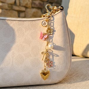 Puede incluir: Un charm dorado con un cierre de mosquetón, sujeto a un bolso de color claro. El charm presenta un colgante con la letra "B", una mariposa rosa, una perla, un lazo, una flor y un relicario en forma de corazón.