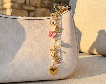 May include: A gold-colored charm with a lobster clasp, attached to a light-colored purse. The charm features a letter "B" pendant, a pink butterfly, a pearl, a bow, a flower, and a heart-shaped locket.