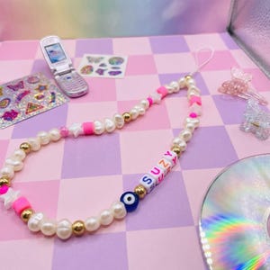 May include: A beaded phone charm featuring white pearls, gold beads, pink accents, and letter beads spelling "SUZY". Includes a blue evil eye bead and a white cord. A flip phone, stickers, and hair clips are in the background.