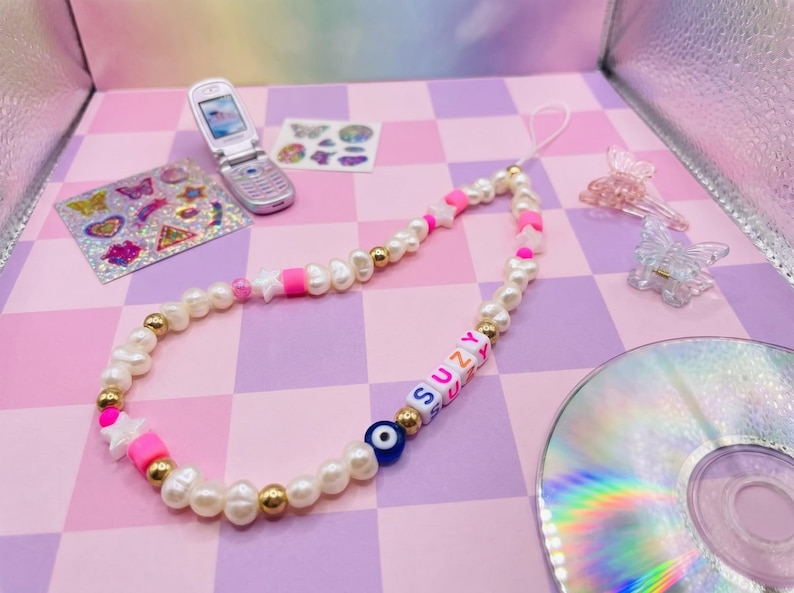 May include: A beaded phone charm featuring white pearls, gold beads, pink accents, and letter beads spelling "SUZY". Includes a blue evil eye bead and a white cord. A flip phone, stickers, and hair clips are in the background.