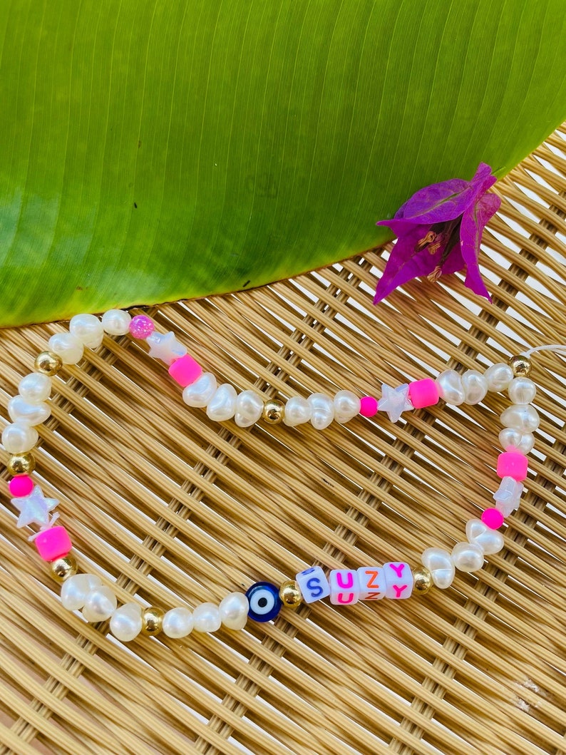 May include: A bracelet with the name "SUZY" spelled out in colourful letter beads. The bracelet features white pearl beads, gold accents, pink and white star-shaped beads, and a blue evil eye charm. The bracelet is displayed on a woven surface.