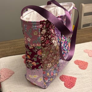 May include: A patchwork tote bag featuring floral patterns in purple, pink, and burgundy. It has a solid purple handle and a white interior. The bag is on a white surface with pink heart-shaped decorations. The bag's dimensions are not visible.