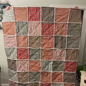 May include: A handmade rag quilt featuring a grid of square fabric patches in various plaid and checkered patterns. The colors include shades of red, black, tan, green, and cream. Each square is joined with exposed, frayed seams in a cross pattern.