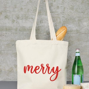 May include: A white canvas tote bag with red embroidered text that says "merry". The bag is filled with a baguette and is sitting on a wooden surface with a bottle of water, a cheese wheel, and a knife.