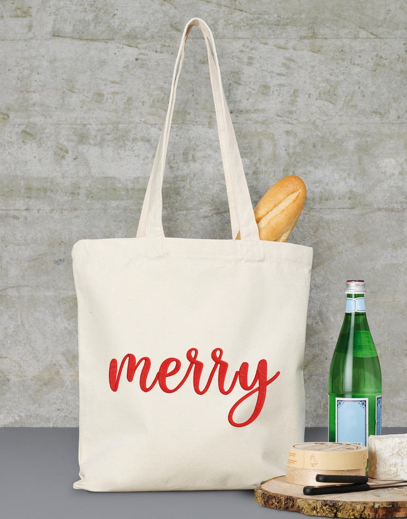 May include: A white canvas tote bag with red embroidered text that says "merry". The bag is filled with a baguette and is sitting on a wooden surface with a bottle of water, a cheese wheel, and a knife.